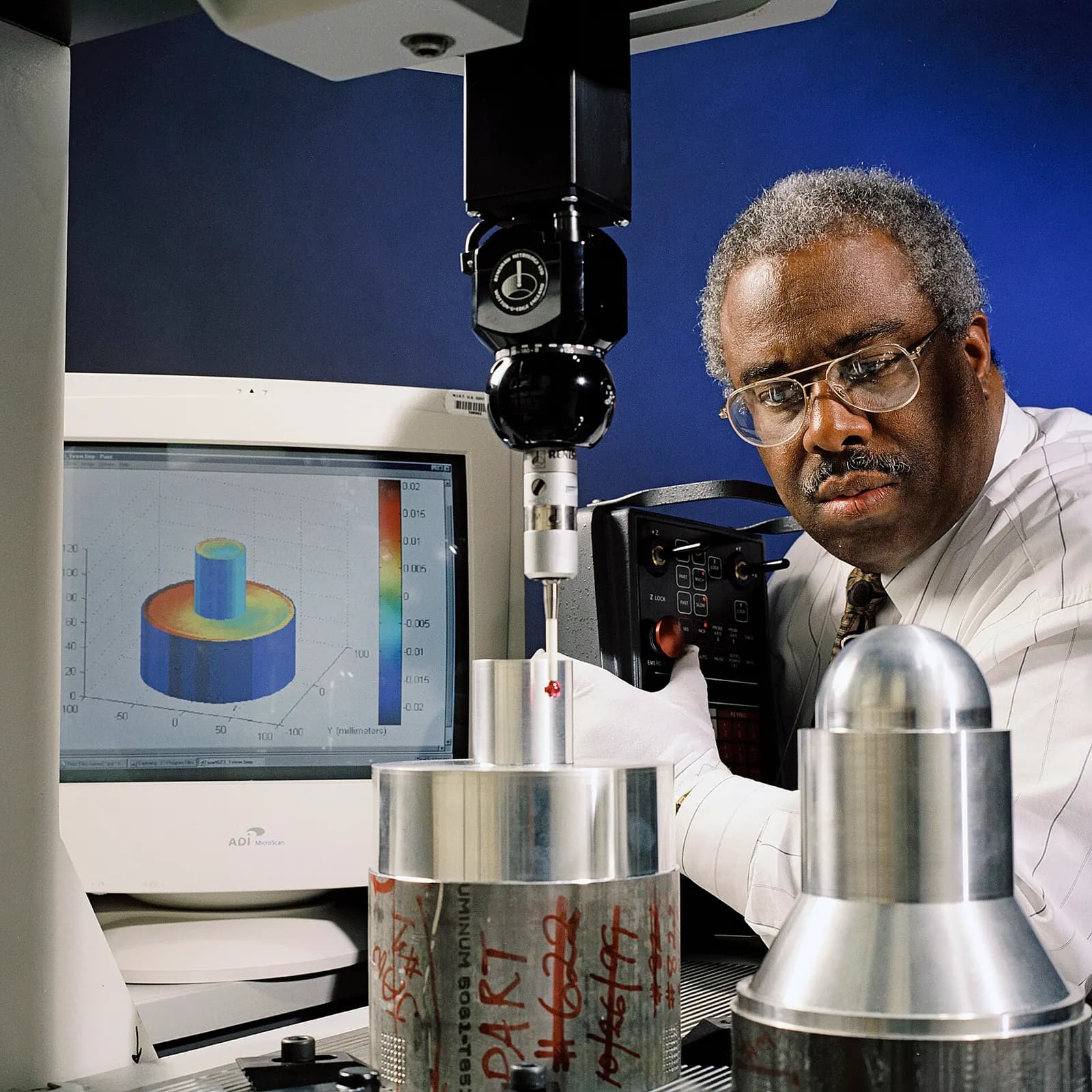 An inspector measuring a machined aluminium part with the probe of a coordinate measuring machine, with the deviation map on screen