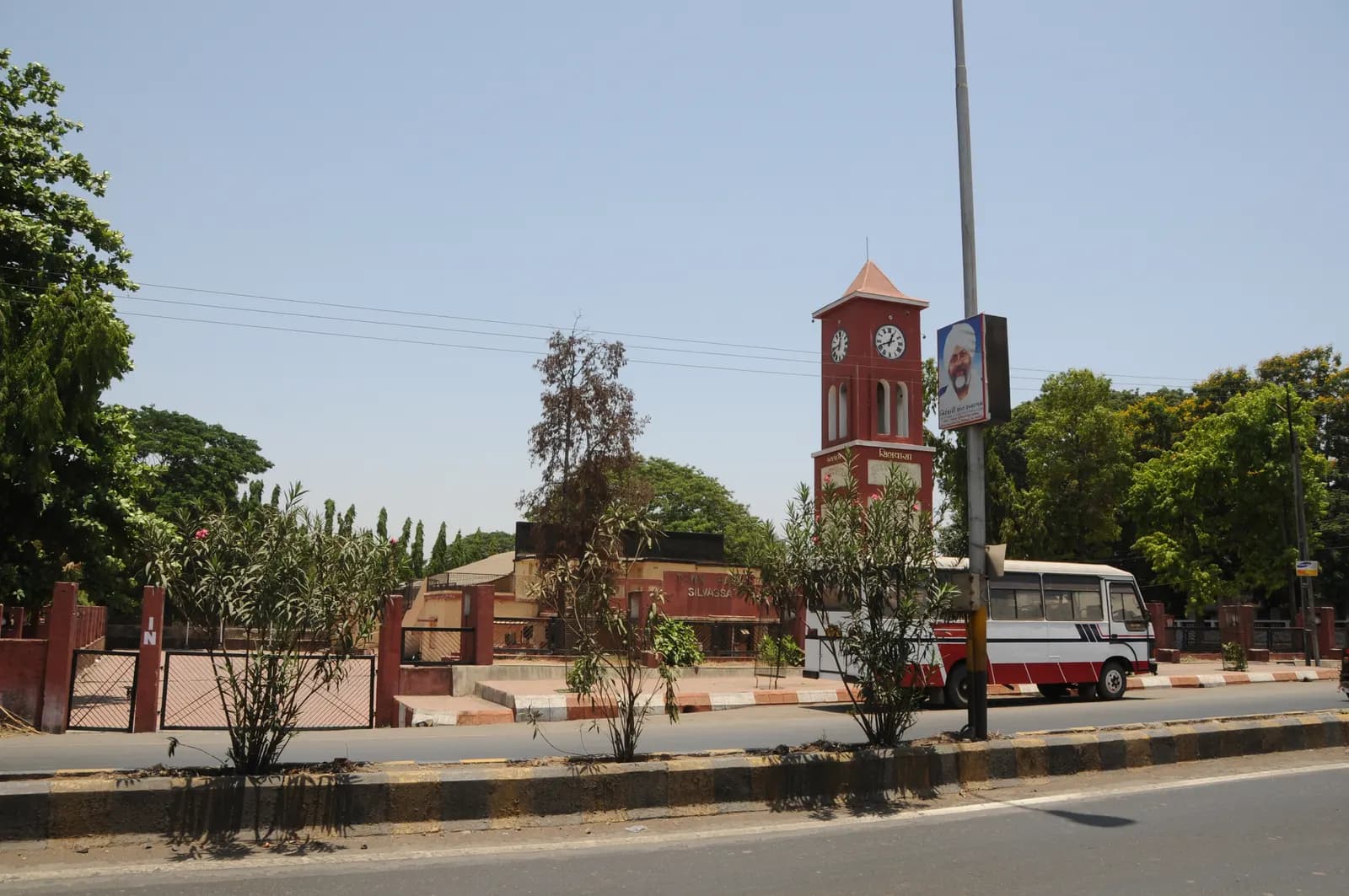 A road in Silvassa with a red clock tower, roadside trees and a passing bus