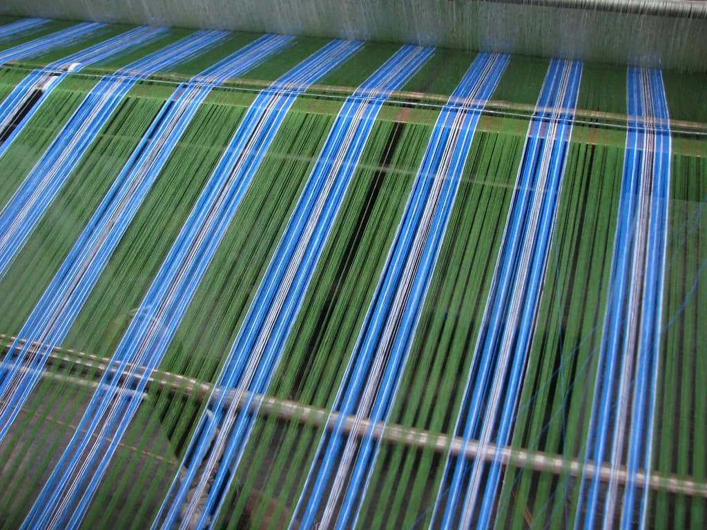 Blue and green cotton warp threads running through a loom as fabric for a lungi is woven