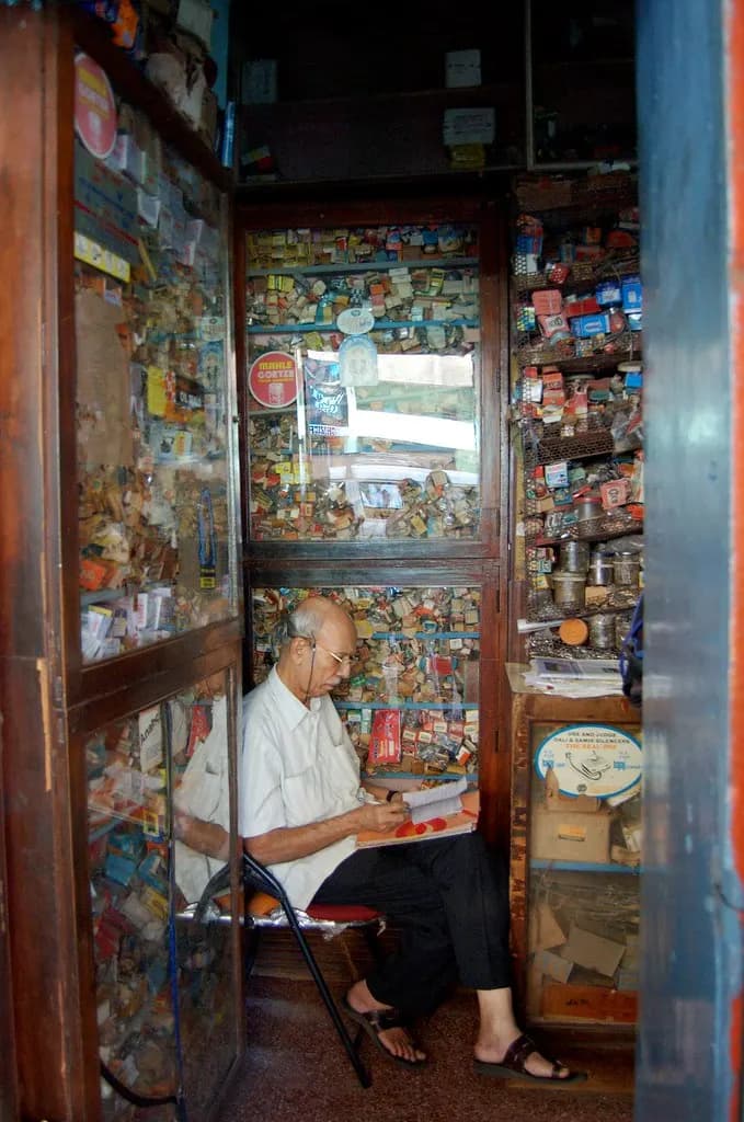 An elderly shopkeeper reading papers inside a small glass-fronted shop crowded with goods