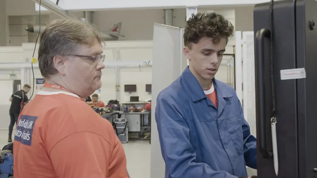 A young trainee in overalls operates a CNC turning machine while an instructor watches at a skills competition