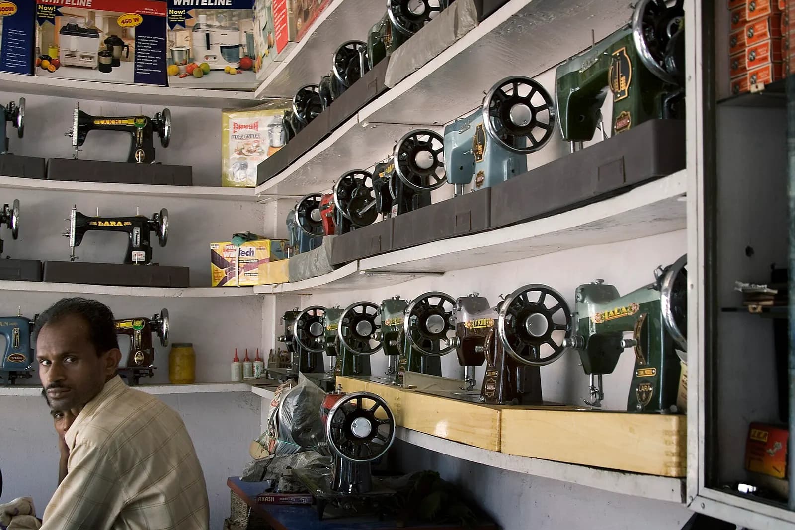 A sewing machine dealer's shop in Varanasi with rows of machines on wall shelves and the owner seated at the counter