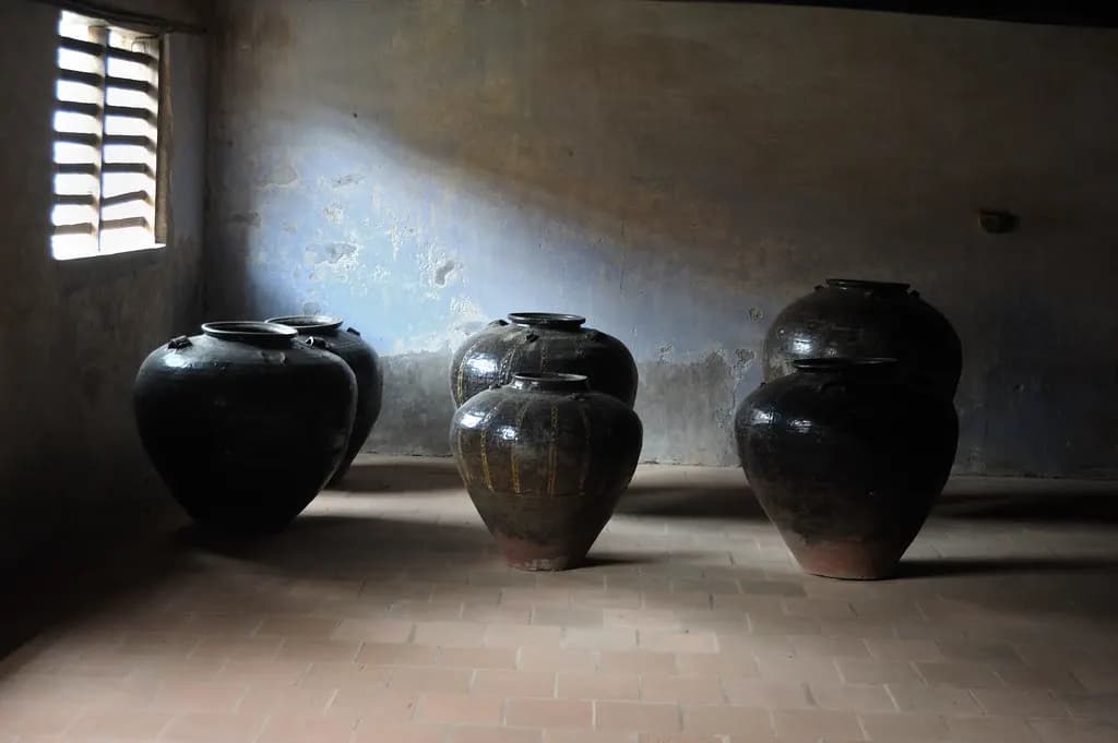 Several large dark glazed earthenware pickle jars standing on the floor of a dim storeroom with a barred window