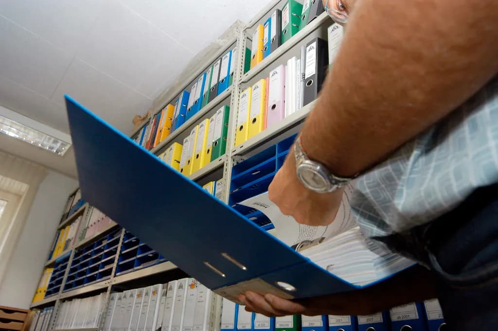 A person flipping through a ring binder in a room lined with shelves of document files