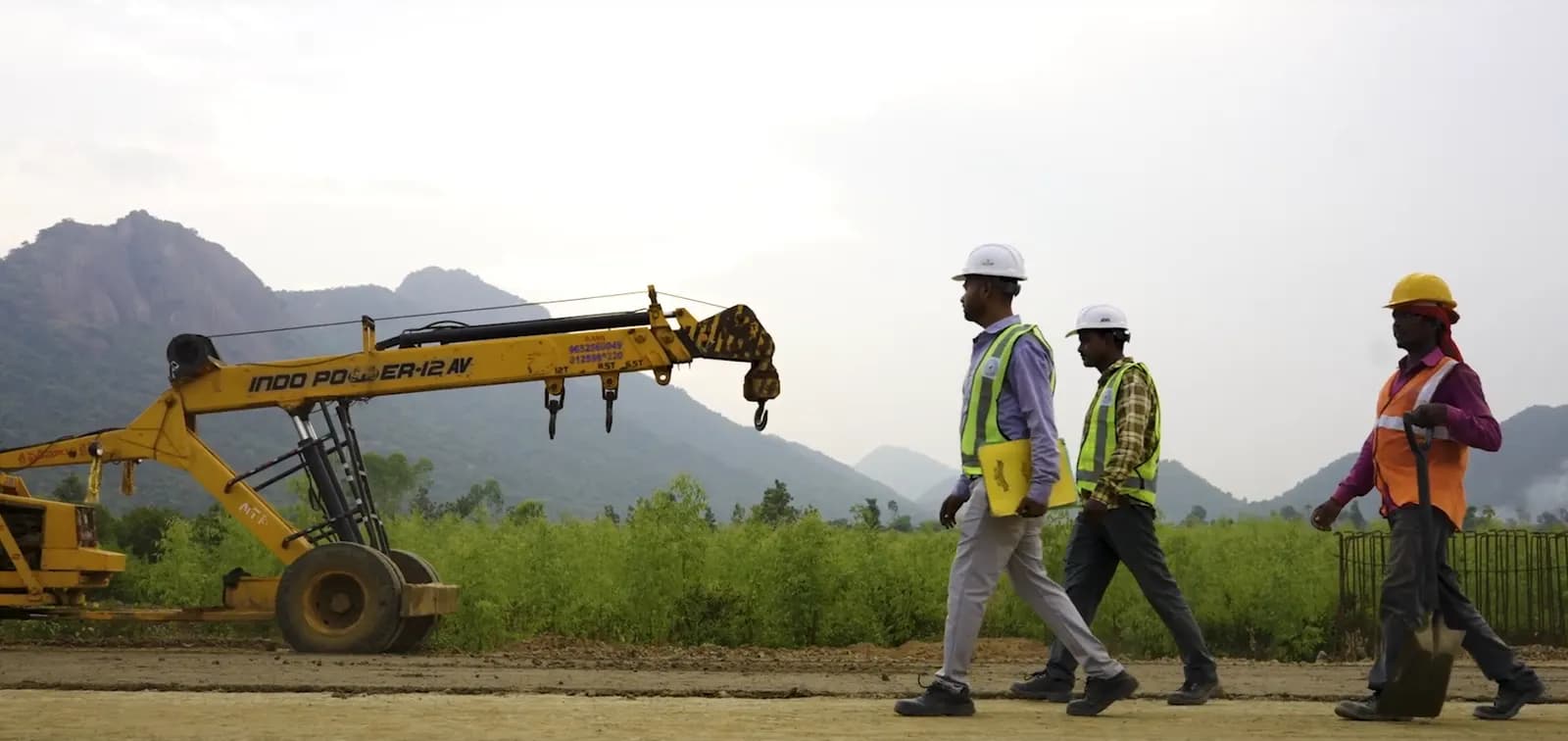 Site engineers in hard hats and safety vests walking past a mobile crane on a project site in Andhra Pradesh