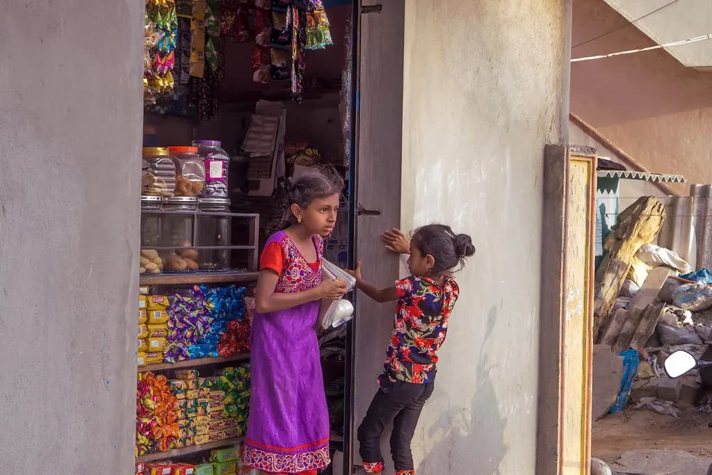 Two girls at the doorway of a small kirana store stocked with packaged snacks and sweets