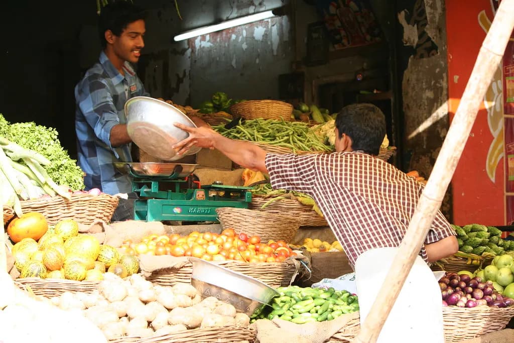A vegetable seller weighing produce for a customer at a market stall in India, with baskets of tomatoes, potatoes and beans