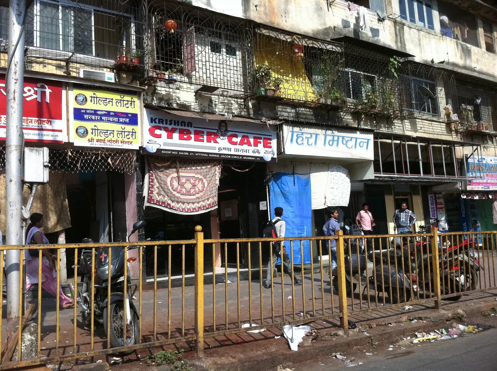 A row of small shops on a Mumbai street — a laundry, a cyber cafe and a sweet shop — with motorcycles parked behind a railing