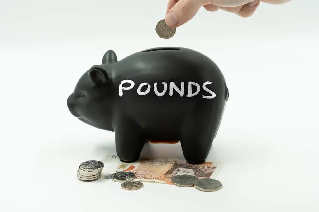 A hand dropping a coin into a black piggy bank labelled POUNDS, with British coins and banknotes beside it