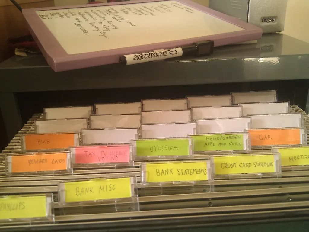 A drawer of labelled hanging files for bank statements, utilities and other papers, below a whiteboard to-do list