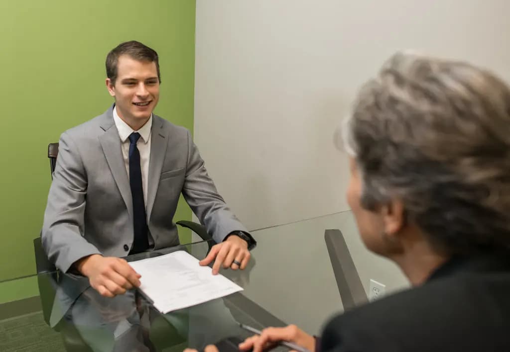 A candidate in a suit sitting across a glass table from an interviewer with paperwork in front of him