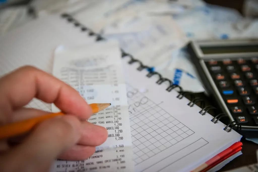 Hand holding a pencil over a till receipt and squared notebook next to a calculator