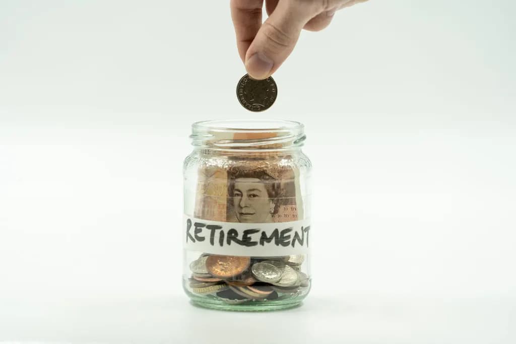 A hand dropping a pound coin into a jar labelled Retirement that holds banknotes and coins