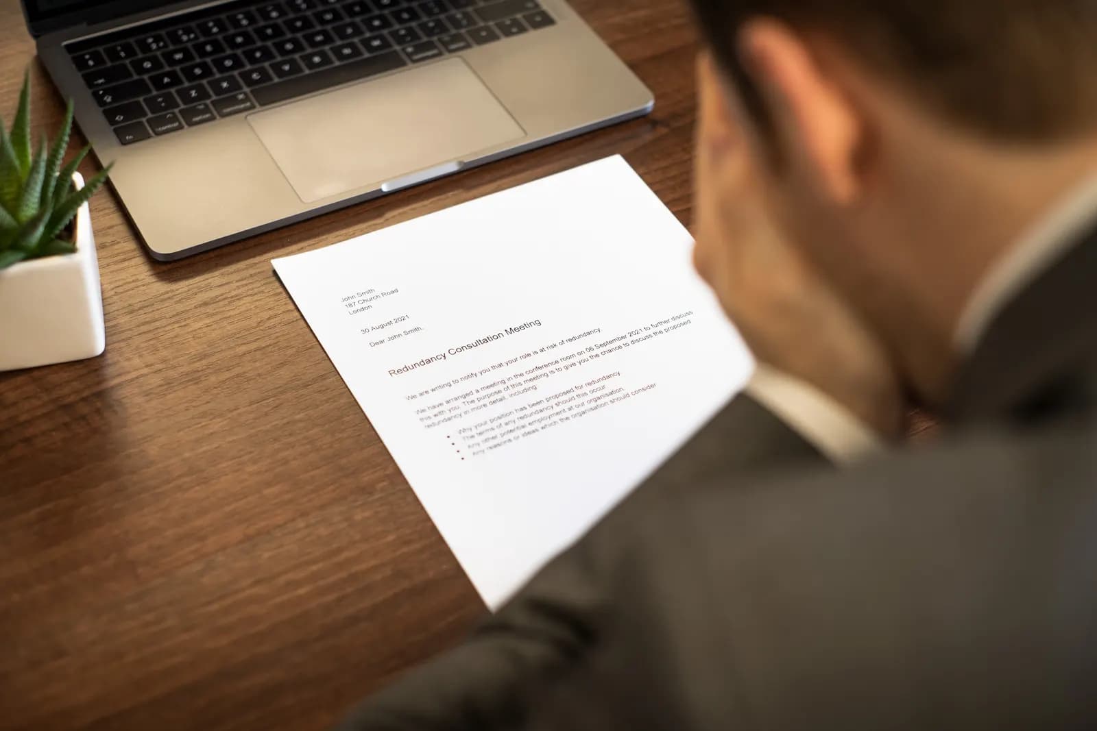 A man reading a letter headed Redundancy Consultation Meeting at a desk beside a laptop