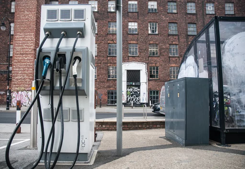 A public electric vehicle rapid charging unit with three cables on a city street with brick buildings behind