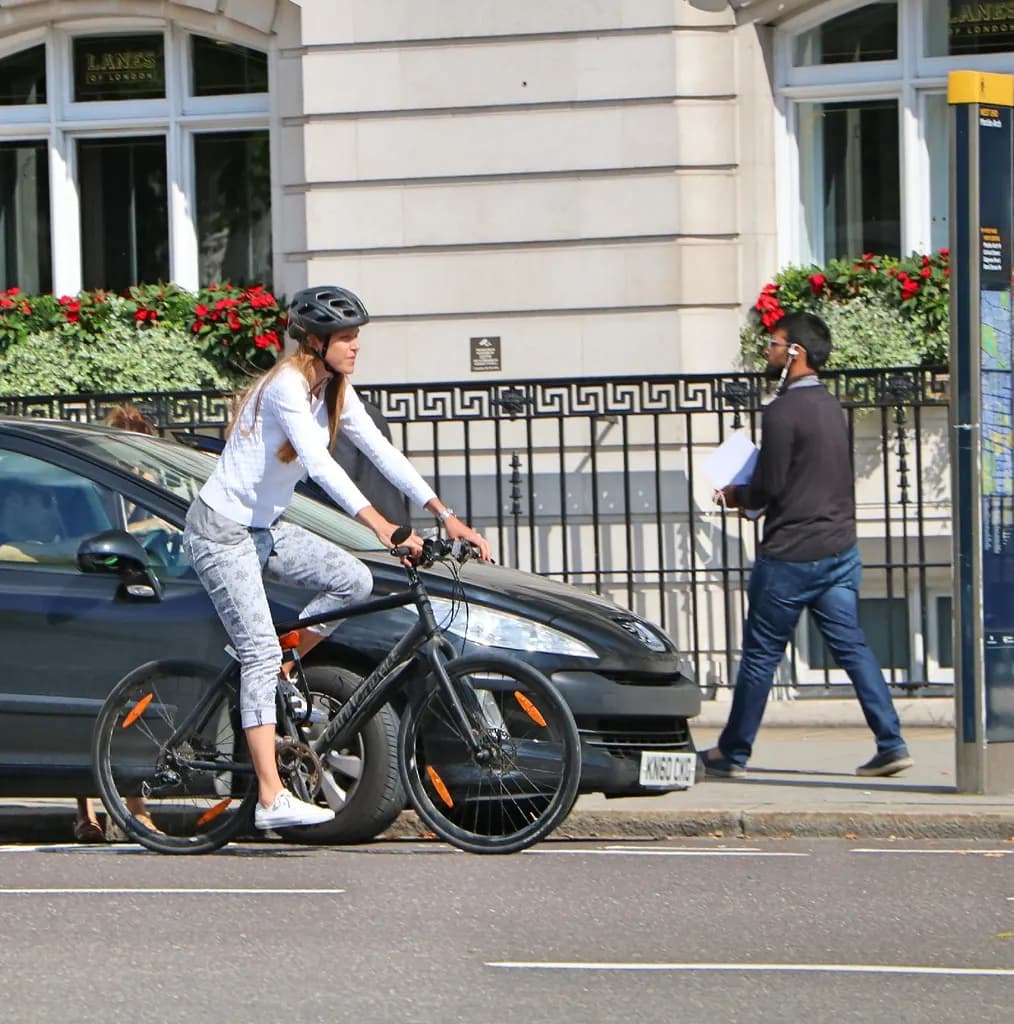 A cyclist in a helmet riding a black hybrid bike along a London street past railings and window boxes