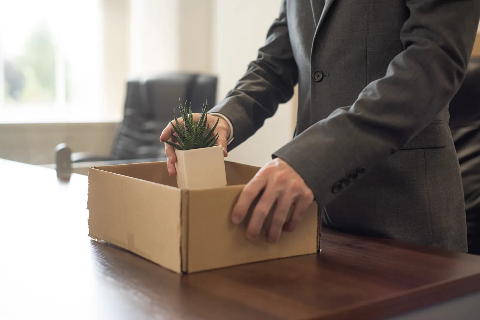 A man in a suit placing a small potted plant into a cardboard box on an office desk