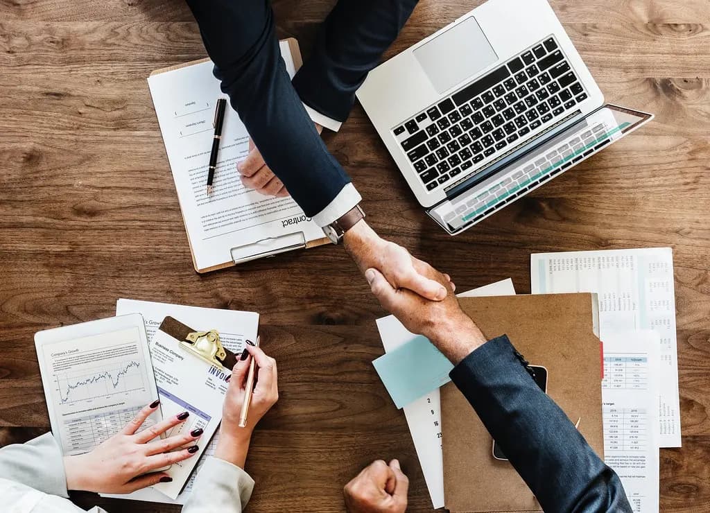 Two people shaking hands across a desk covered with financial papers, a laptop and a clipboard