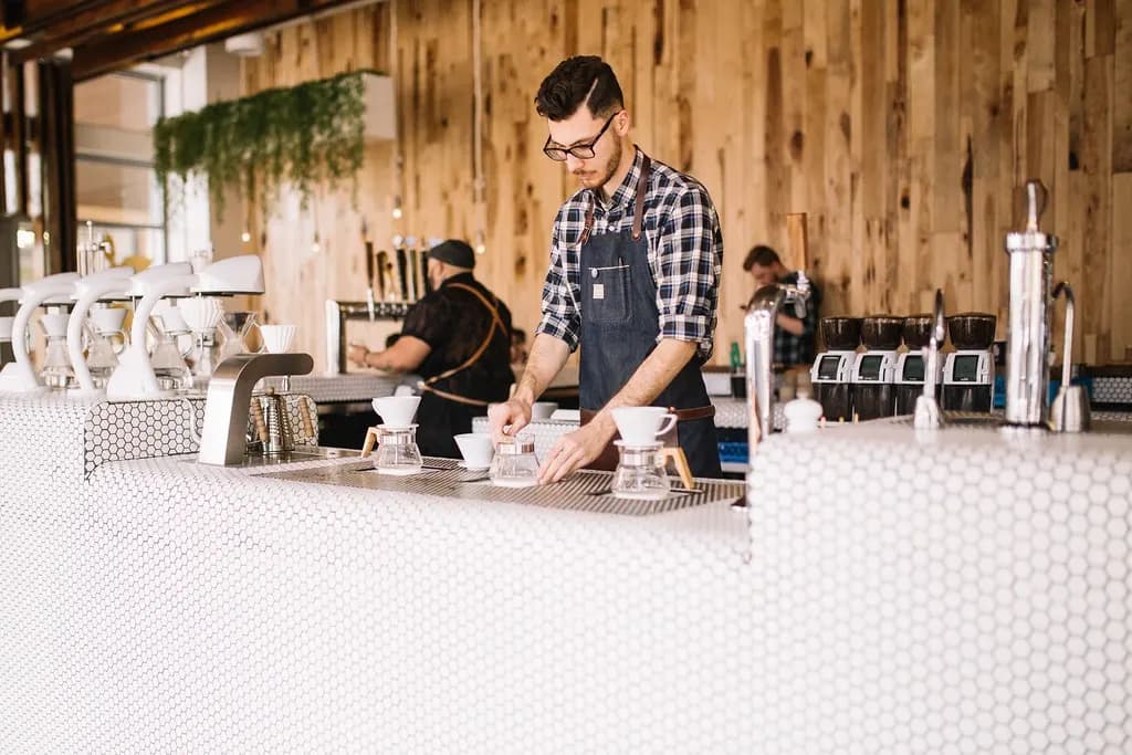 Barista in an apron preparing pour-over coffee behind a cafe counter while coworkers work in the background