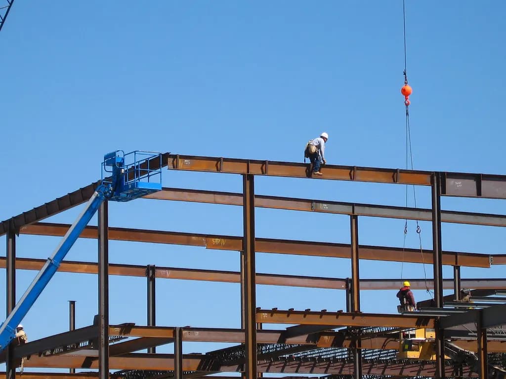 Ironworker standing on the steel frame of a building under construction against a blue sky, with a boom lift alongside