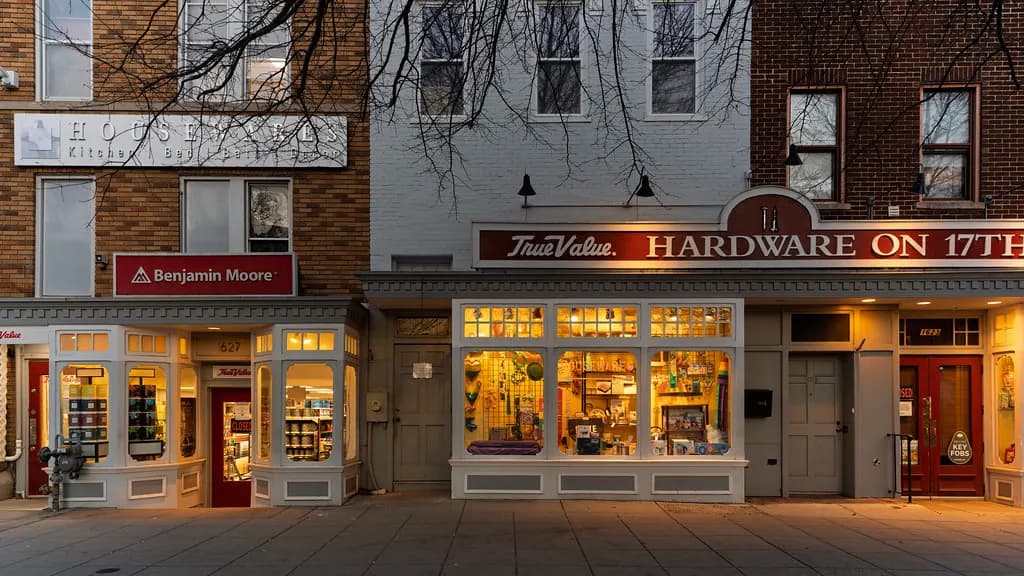 Row of shopfronts at dusk with a hardware store signed 'Hardware on 17th' next to a paint and kitchenware shop