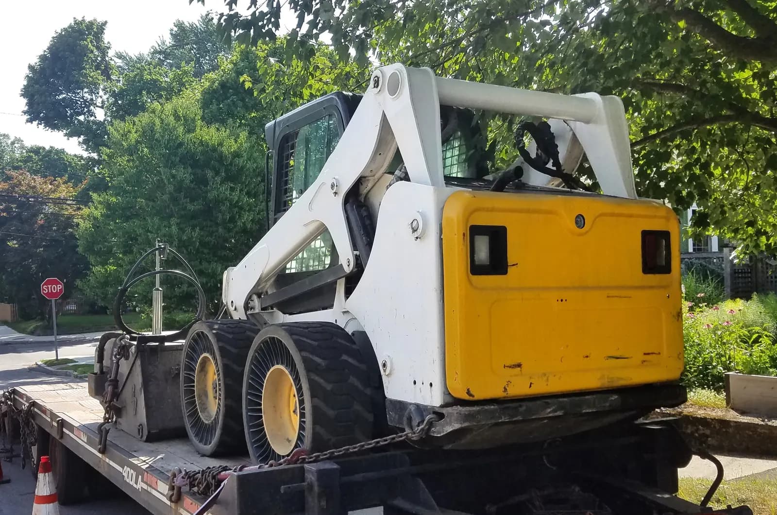 A compact skid steer loader chained onto a flatbed trailer parked at the curb