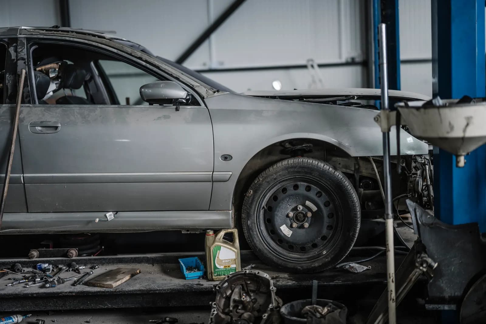Partially disassembled silver car raised on a lift in an auto repair shop with tools and parts on the floor