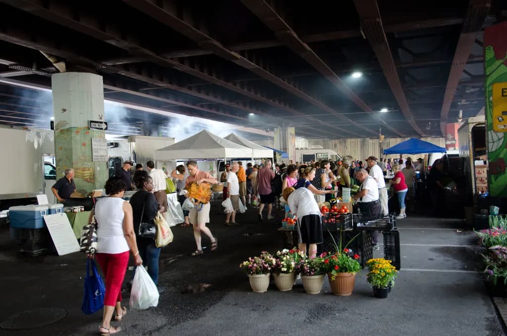 Shoppers walk between vendor tents and flower displays at a farmers market under a highway overpass in Baltimore