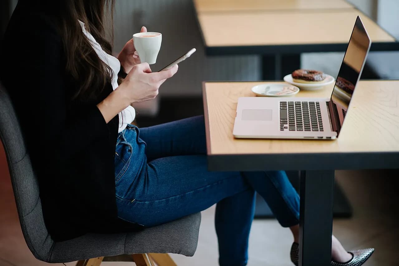 A woman in a cafe holds a coffee cup and scrolls on her smartphone beside an open laptop