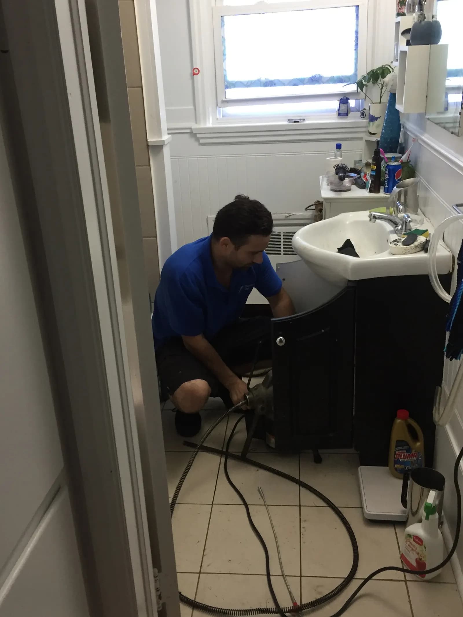 Plumber kneeling under a bathroom sink with a drain-cleaning machine and hoses on a tiled floor