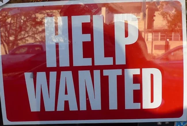 Red and white Help Wanted sign taped inside a shop window, with street reflections in the glass