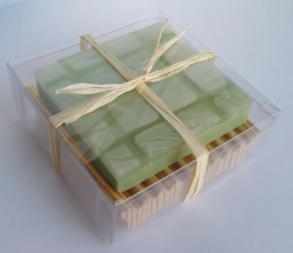 A green handmade soap bar on a bamboo soap dish, tied with raffia inside a clear gift box