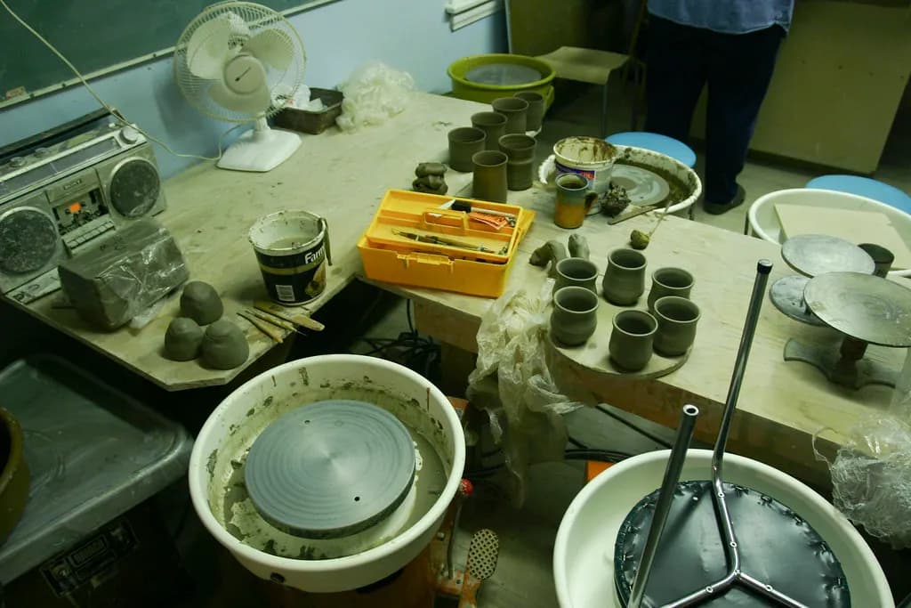 A pottery studio workbench with freshly thrown clay cups, glaze buckets, tools and two potter's wheels