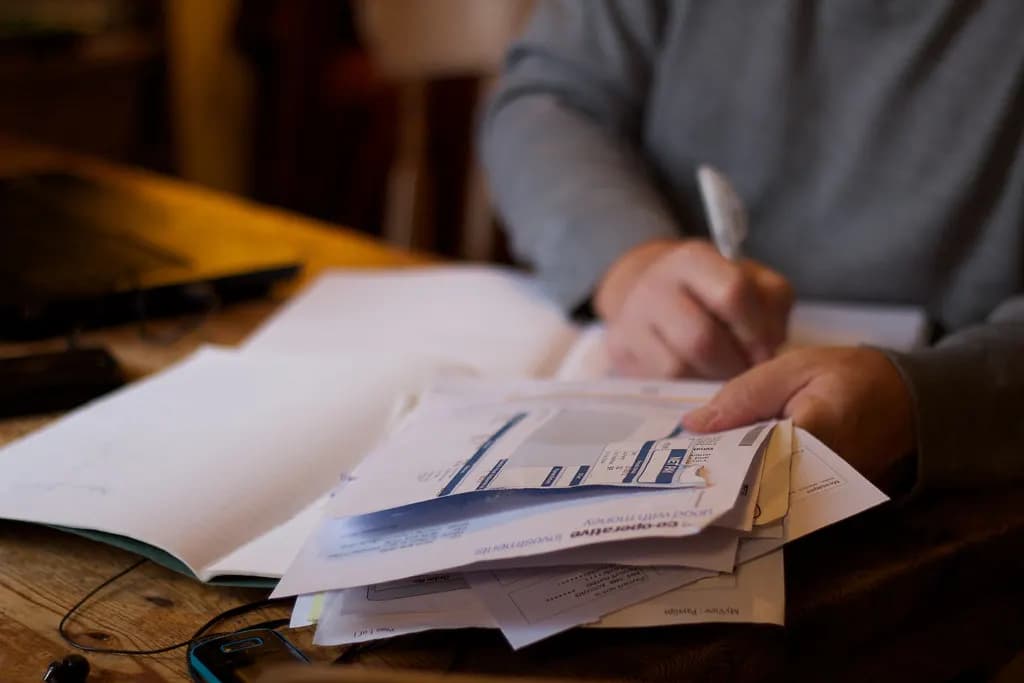 A person at a kitchen table sorting a pile of paper bills and receipts with a pen and notebook