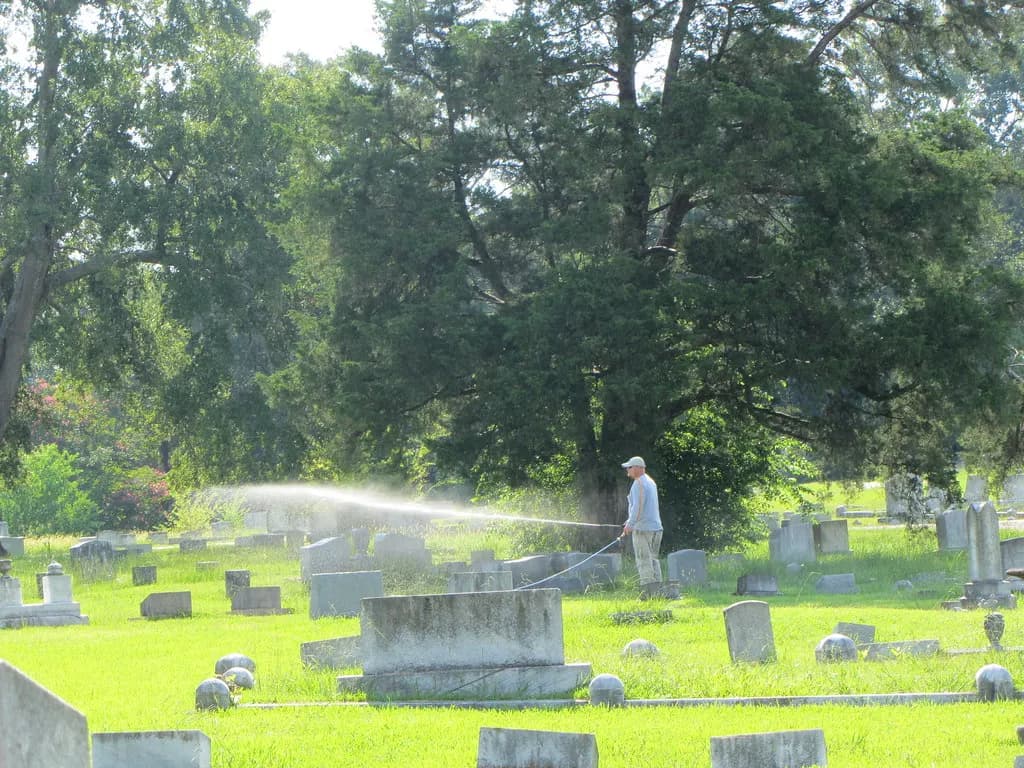 A worker spraying herbicide with a hose across a large grassy cemetery lawn