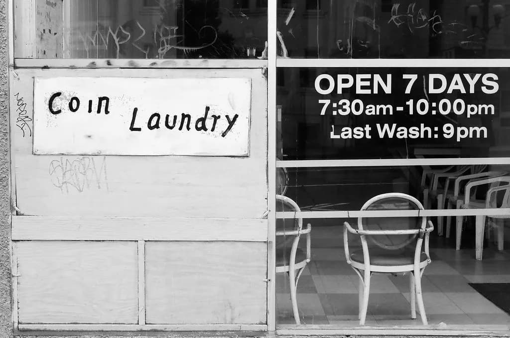Storefront of a coin laundry with a hand-painted Coin Laundry sign and window lettering listing open 7 days, 7:30am to 10:00pm, last wash 9pm
