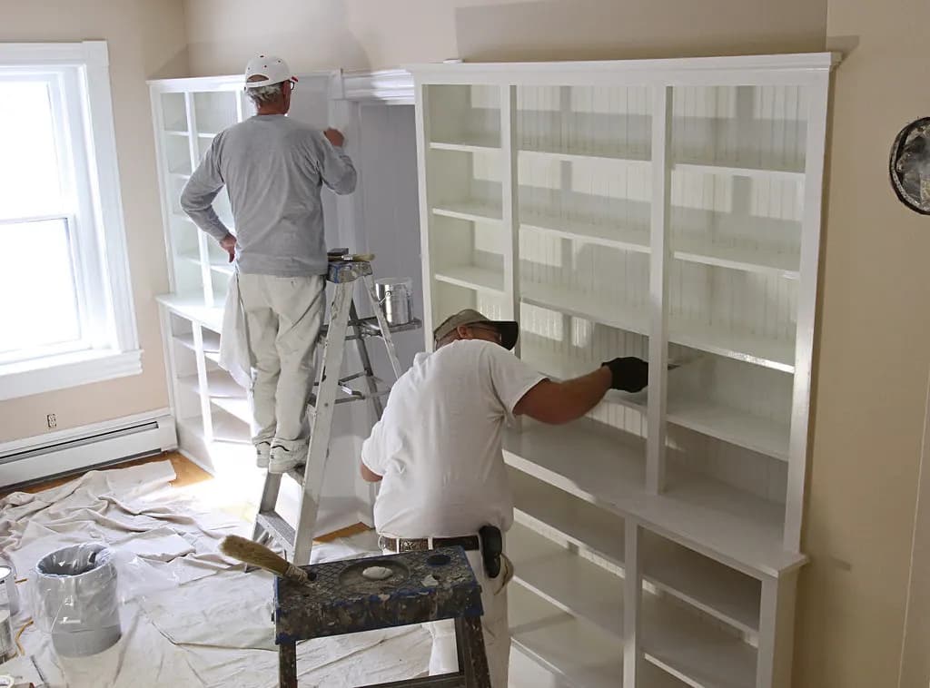 Two painters on step ladders painting built-in white shelving in a room with drop cloths on the floor