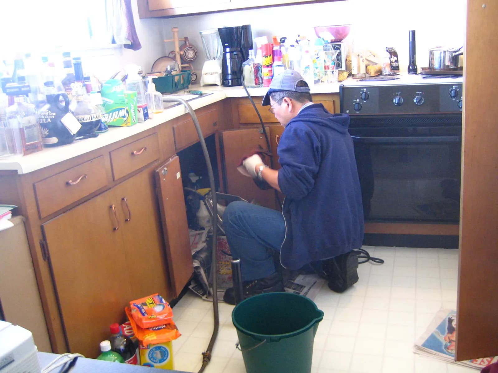 A plumber kneeling at an open kitchen sink cabinet, working on the drain with a drain-cleaning cable and a bucket beside him