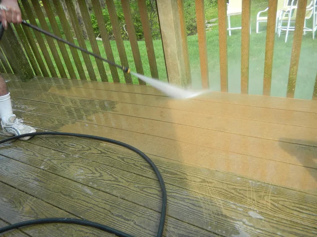 A pressure washer wand spraying a weathered wooden deck, leaving a clean stripe across the boards
