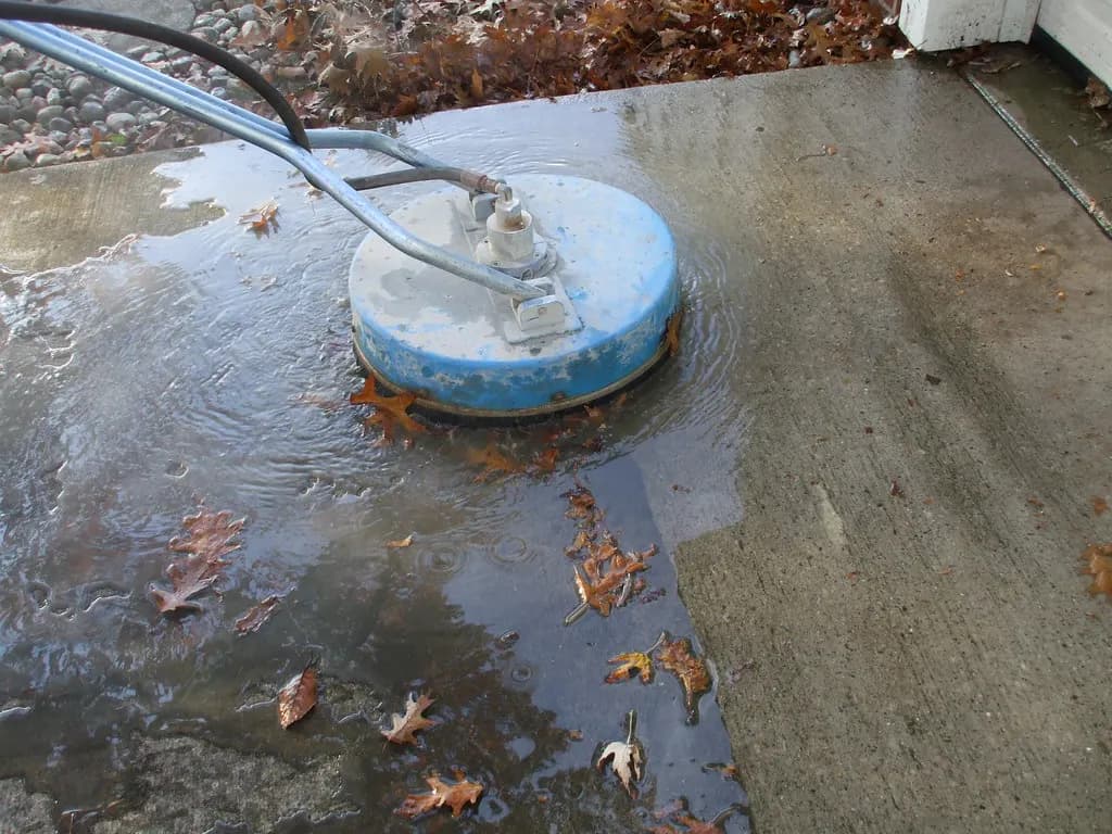 A rotary surface cleaner attachment cleaning a concrete walkway, with dirty water and leaves running off the slab