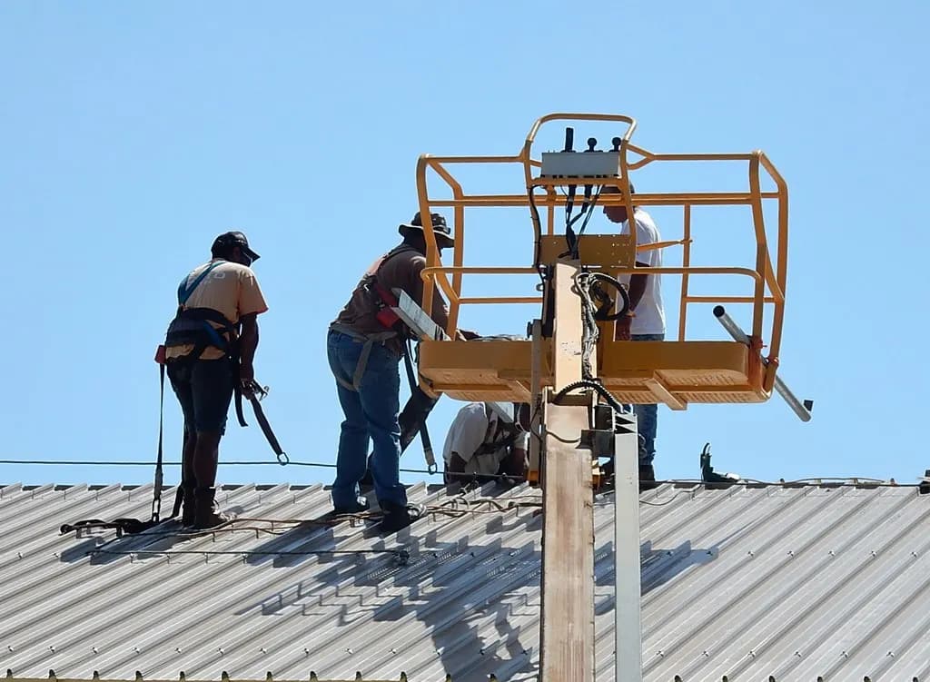 Two roofers wearing fall-arrest harnesses on a metal roof beside an aerial lift