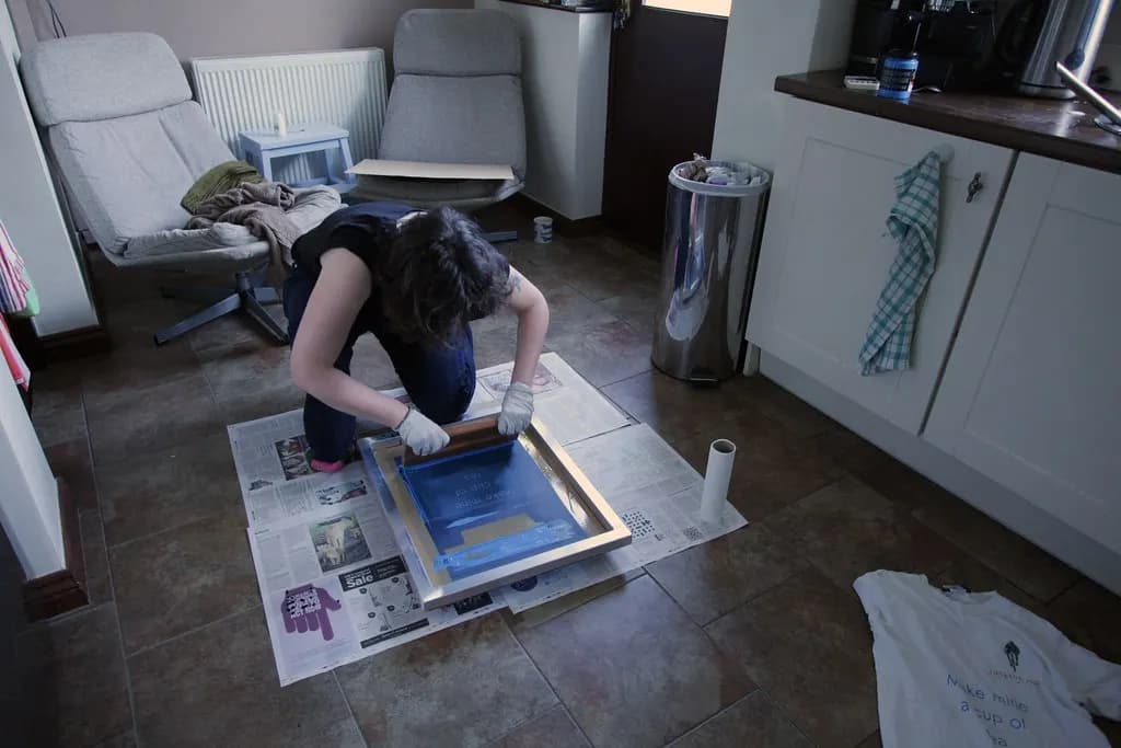 A person kneeling on newspaper at home pulling ink through a screen-printing frame onto a T-shirt