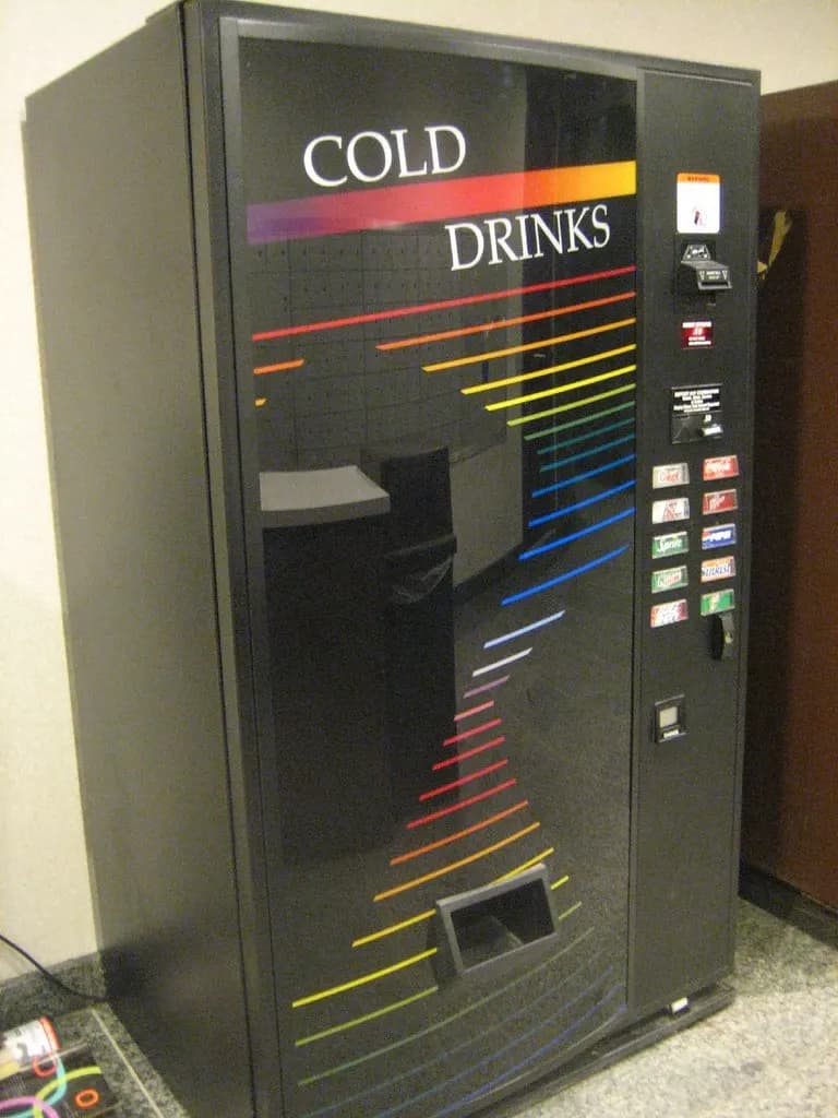 A black cold-drinks vending machine with selection buttons, a bill acceptor and a coin slot standing against an office wall