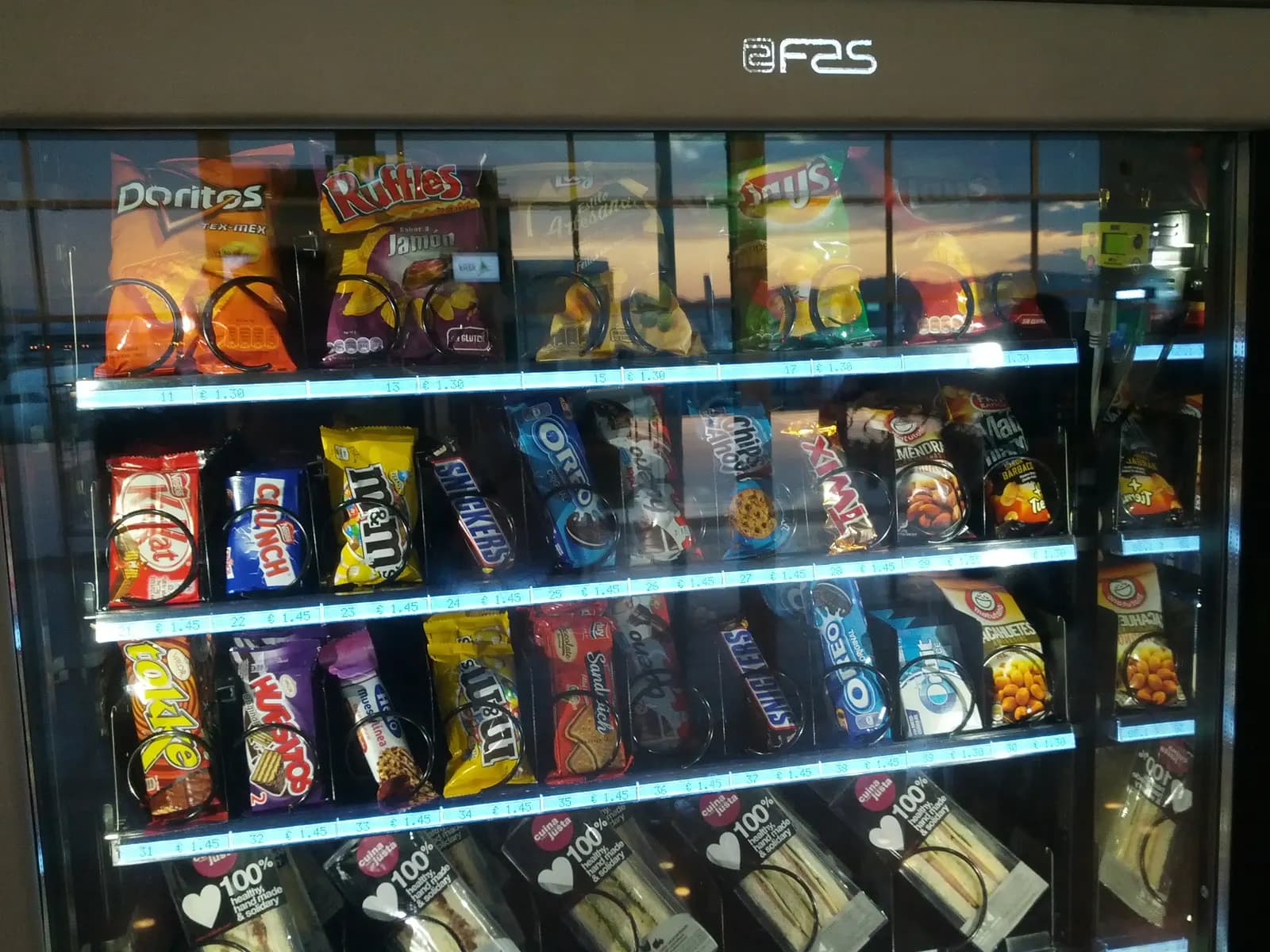 A glass-front snack vending machine with chips, candy bars, cookies and sandwiches in spiral coils, each slot labelled with a price