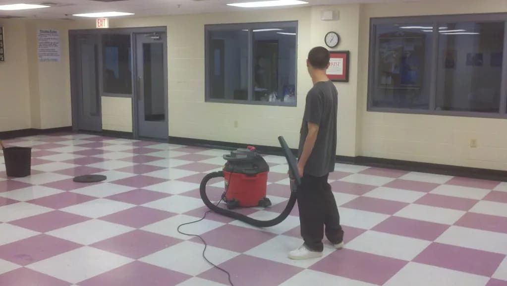 A janitor uses a red wet-dry vacuum on a checkered tile floor in an empty commercial building hallway