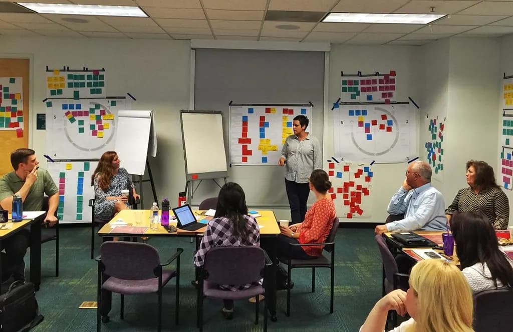 A consultant stands at the front of a meeting room facilitating a workshop, with walls covered in sticky-note planning boards and participants seated around tables