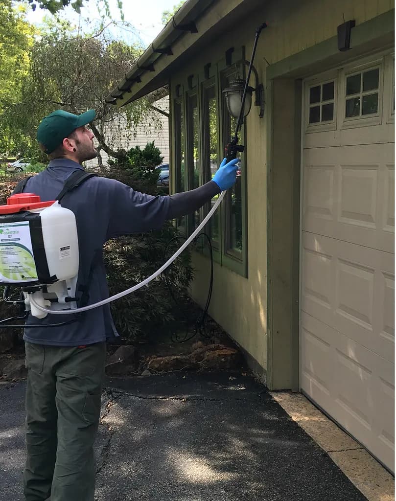 A pest control technician wearing gloves and a backpack sprayer treats the eaves and siding of a house beside a garage door