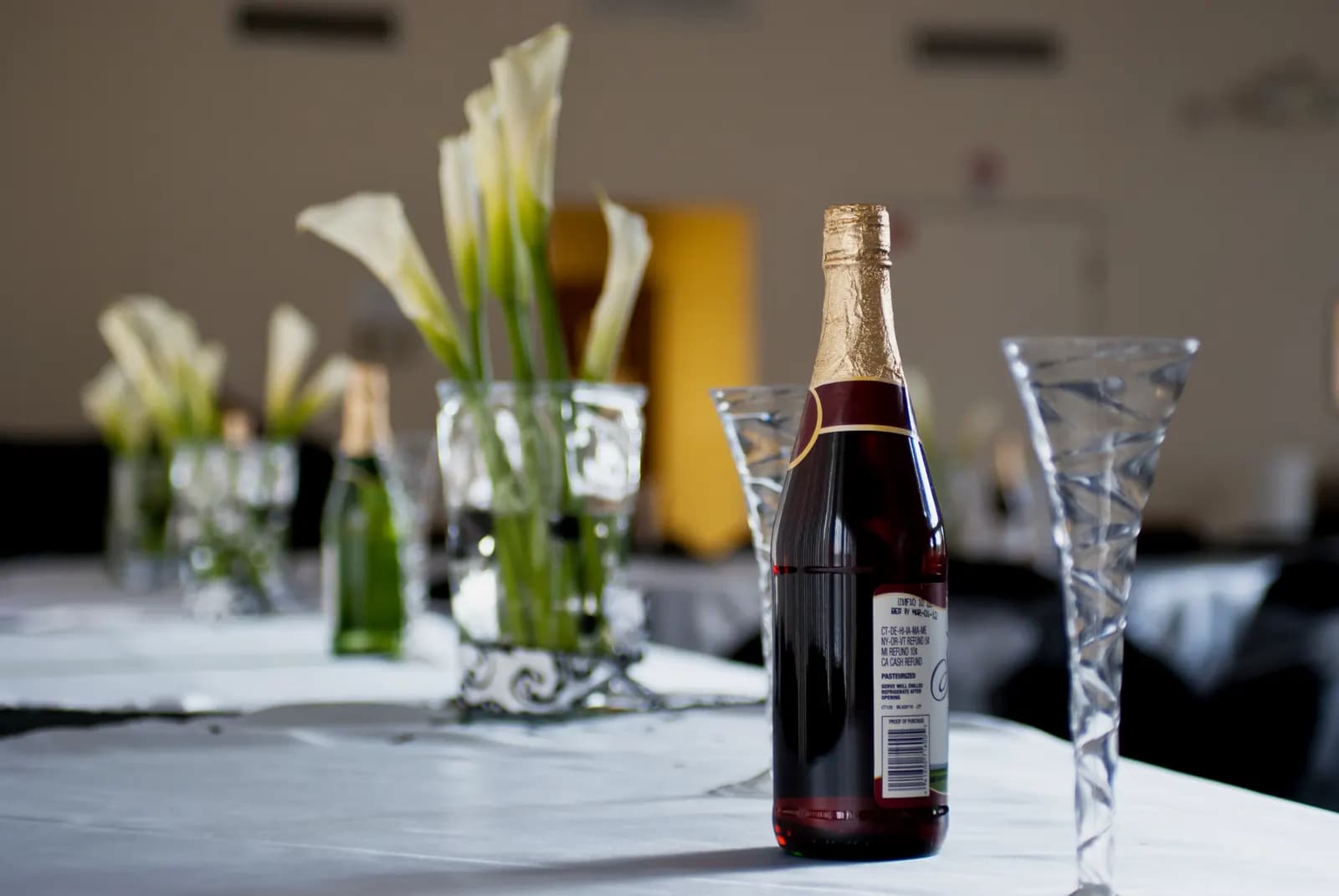 A wedding reception table with white linen, calla lily centerpieces, champagne flutes and a bottle of sparkling wine