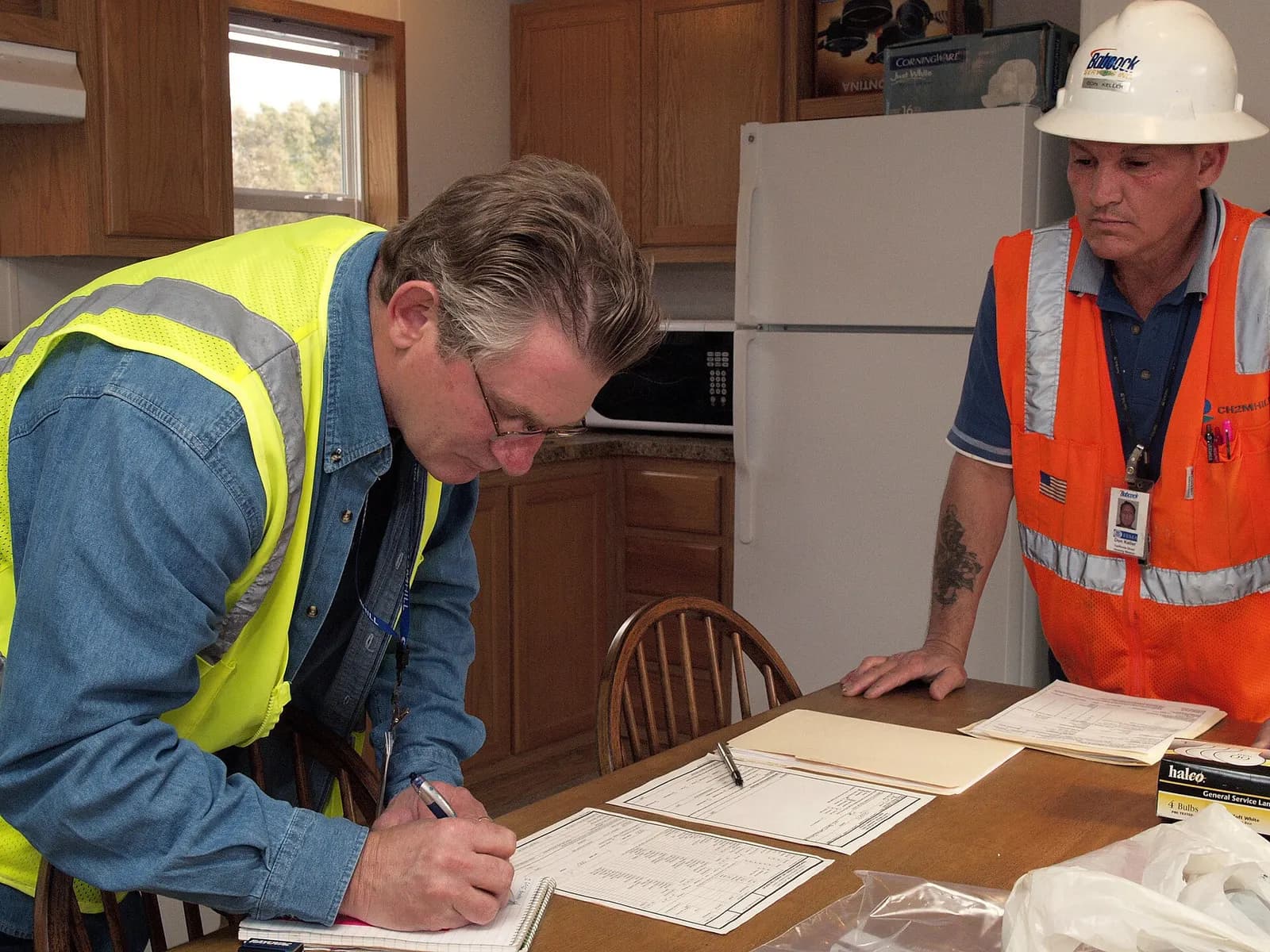 Contractor in a high-visibility vest signing job paperwork at a kitchen table while a colleague in a hard hat looks on