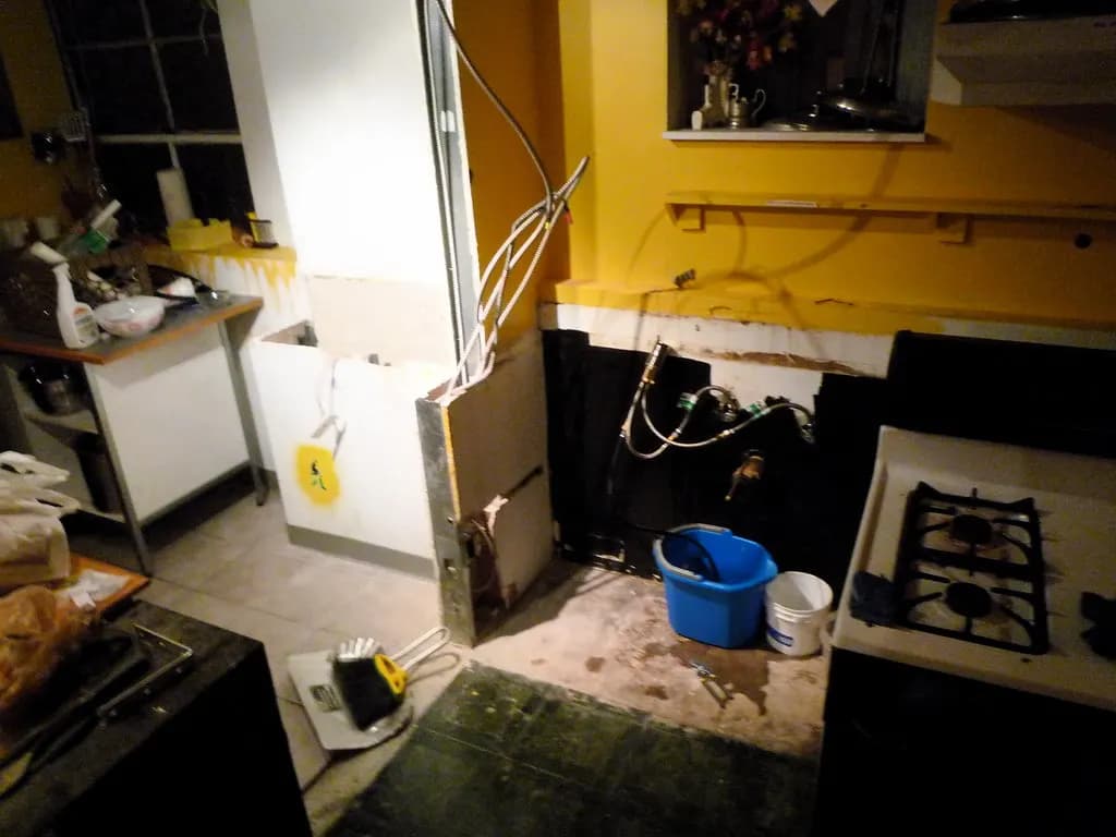 Kitchen mid-renovation with cabinets removed, exposed plumbing lines and a bucket under the sink connections